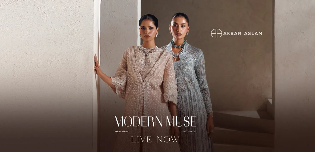 Two models wearing elegant, long-sleeved traditional outfits by Akbar Aslam, standing in a doorway. The model on the left wears a light beige textured kurta with a matching dupatta, while the model on the right wears a shimmering silver-gray kurta.