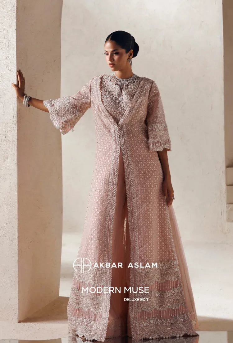 A woman models a light pink embroidered gown with a matching sheer jacket in an elegant indoor setting with beige columns. The brand text 'AKBAR ASLAM' and 'MODERN MUSE DELUXE EDIT' is displayed at the bottom.