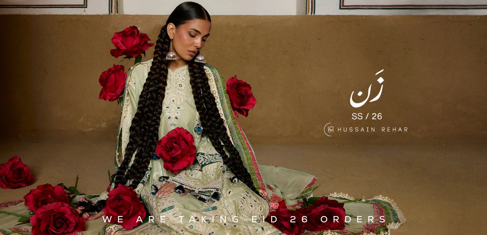Woman in traditional Pakistani outfit with red flowers, sitting on a brown couch with 'Hussain Behar' branding Eid Dresses.