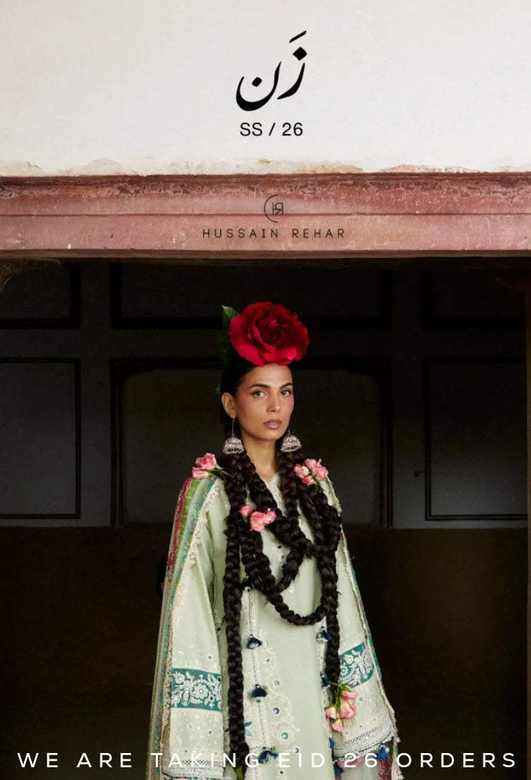 Woman in traditional Pakistani outfit with red flowers, sitting on a brown couch with 'Hussain Behar' branding Eid Dresses UK.