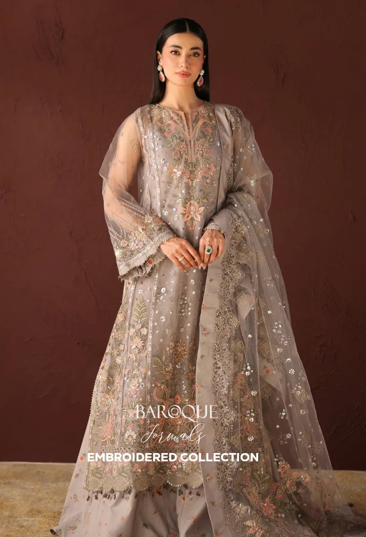 A woman models a Baroque Jewels embroidered traditional outfit in a sheer taupe-gray tulle with intricate floral embroidery in gold, pink, and green, set against a dark brown textured background.