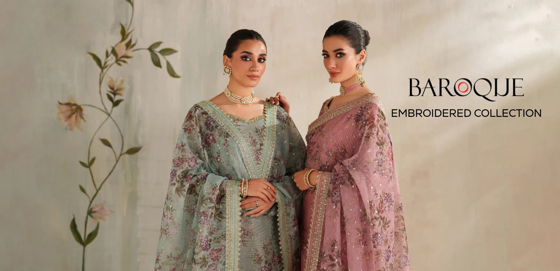 Two women modeling traditional embroidered ethnic outfits against a soft beige wall with floral artwork. The left model wears a light blue-green embroidered kurta set, and the right model wears a dusty pink embroidered kurta set.