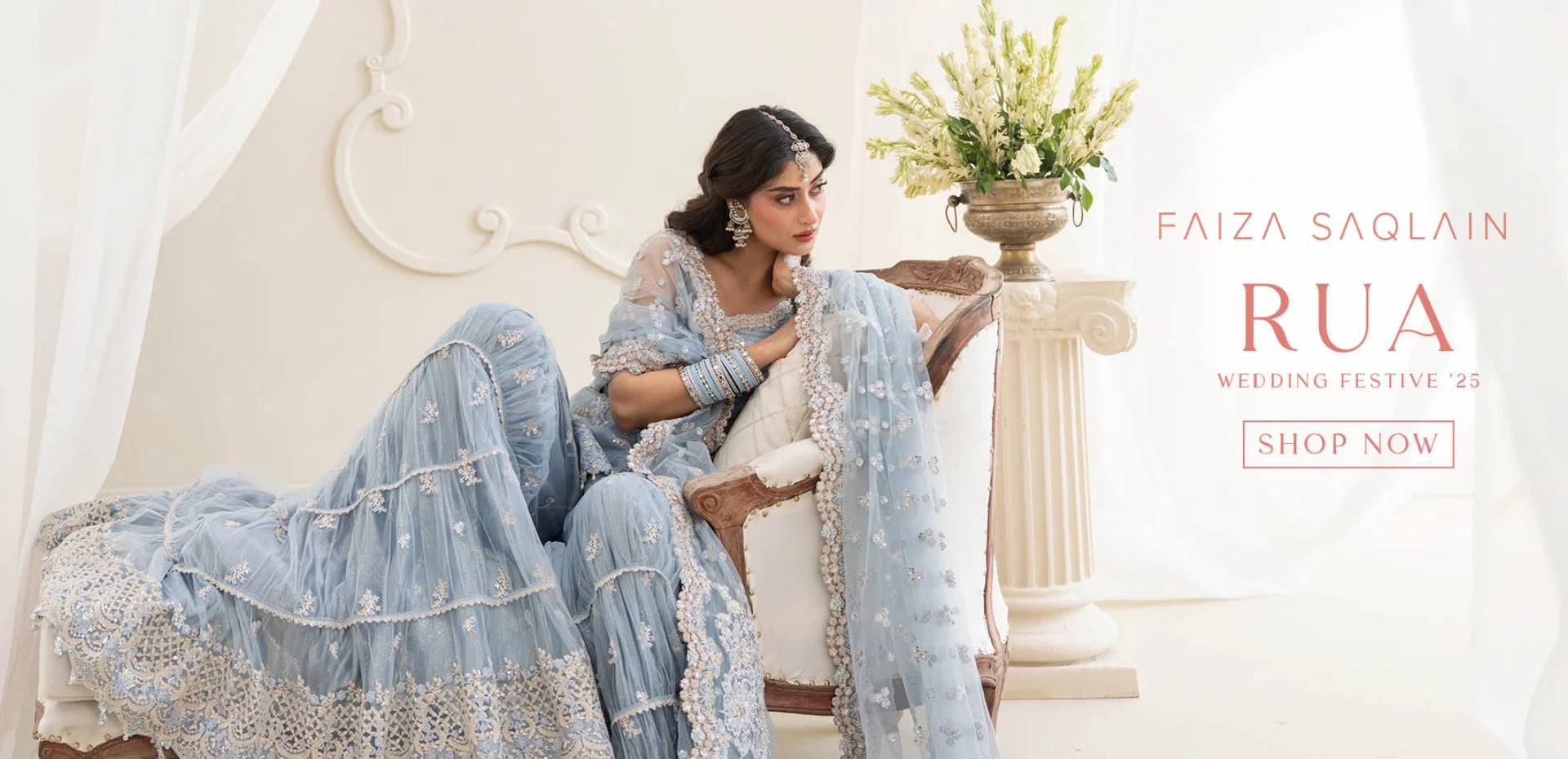 Woman in a light blue embroidered outfit sitting on a white couch with floral decorations, Faiza Saqlain RUA branding visible.